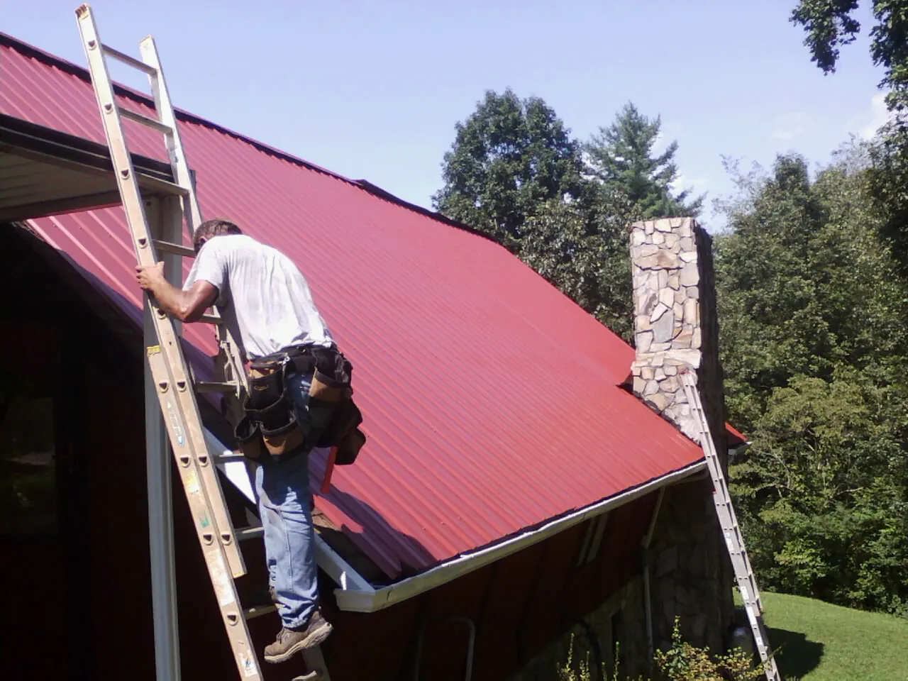 Roofer on a ladder beside a red metal roof and stone chimney — Soffit & Fascia Repair by Roofing Brothers in Fort Recovery
